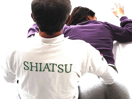 Woman in purple bathrobe on lounger, behind her woman in white with Shiatsu logo massaging